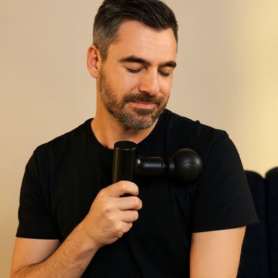 Un homme masse son bras avec le pistolet massant - RecoveryPro dans le confort de son canapé